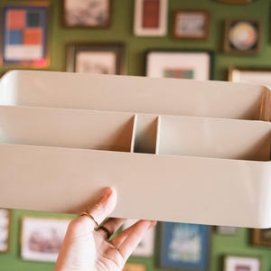 May include: A beige rectangular organizer with three compartments is held up. The organizer has rounded edges and is made of a smooth, solid material. The background features a green wall with framed artwork.