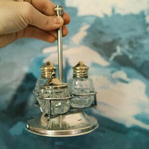 Puede incluir: Un juego de salero y pimentero vintage. El juego presenta tres botellas de vidrio transparente con un patrón de diamantes, sujetas en un marco de metal plateado. Las tapas son de color dorado. El juego es sostenido por una mano.