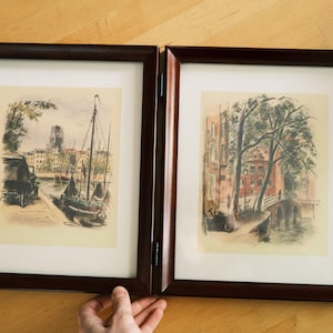 May include: A hinged double picture frame made of dark brown wood, containing two separate watercolor-style illustrations. The left print depicts a harbor scene with a sailboat, while the right print shows a canal scene with trees and a bridge.