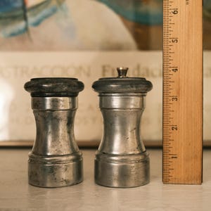 May include: Two antique silver salt and pepper shakers with dark gray tops. The shakers have a classic hourglass shape and are next to a wooden ruler. The shakers are approximately 3 inches tall.