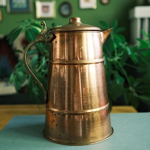 May include: A tall, cylindrical copper pitcher with a polished metallic finish, featuring a sturdy curved handle, a hinged lid with a small knob, and a pointed pouring spout. The vessel has a smooth, tapered body with visible horizontal seams.