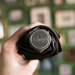 Puede incluir: Un paraguas negro enrollado con una tapa plateada. La tapa tiene la palabra "Kairos" grabada. El paraguas se sostiene en una mano, con un fondo verde borroso.