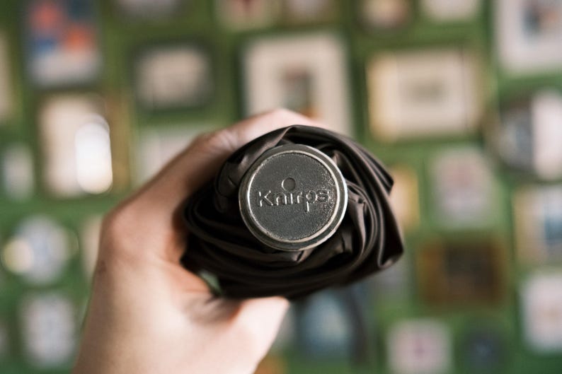 Puede incluir: Un paraguas negro enrollado con una tapa plateada. La tapa tiene la palabra "Kairos" grabada. El paraguas se sostiene en una mano, con un fondo verde borroso.