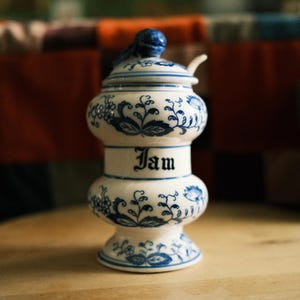 May include: A white ceramic jam jar with a blue floral pattern and a matching lid featuring a small knob. The word Jam is printed in black gothic-style lettering on the center of the jar. The jar has a rounded, hourglass-like shape.