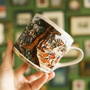 May include: A white ceramic mug with a detailed landscape scene in brown, beige, and blue tones. The mug has a gold rim and a curved handle. The background is a green wall with framed pictures.