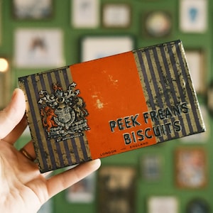 May include: Vintage Peek Freans Biscuits tin. The rectangular tin features a gold and black striped design with an orange center panel. The tin has a crest and the text "Peek Freans Biscuits London England."