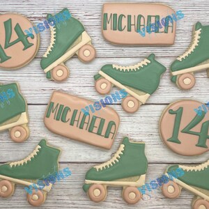 May include: A set of decorated sugar cookies featuring a green and white roller skate design, a pink cookie with the number 14, and two pink cookies with the name "Michaela" written on them.