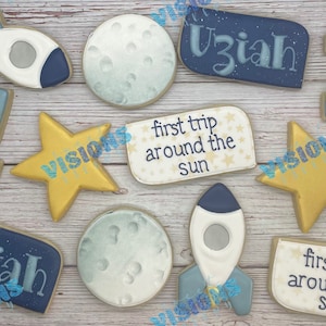 May include: A set of nine decorated sugar cookies with a space theme. The cookies feature a rocket, a moon, a star, a sun, and the words "first trip around the sun" and "Uziah".