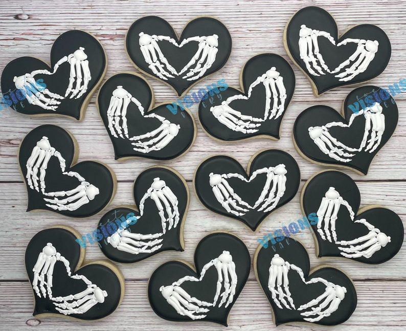 K&ouml;nnte beinhalten: Zw&ouml;lf schwarze herzf&ouml;rmige Kekse, die mit wei&szlig;em Zuckerguss in Form von Skeletth&auml;nden verziert sind. Die Kekse sind in einem Kreis auf einer wei&szlig;en Holzoberfl&auml;che angeordnet.