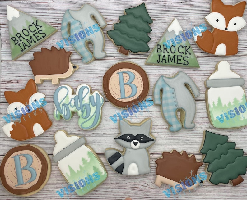 May include: A set of 12 decorated sugar cookies with woodland and baby themes. The cookies feature a fox, a raccoon, a hedgehog, a baby onesie, a baby bottle, a tree, a mountain, and a wood slice with the letter B. The cookies are decorated with blue, green, brown, and white icing.