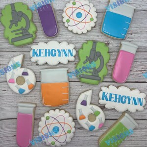 May include: A collection of science-themed sugar cookies featuring gray microscopes on green backgrounds, purple and pink test tubes, white atom shapes with colorful orbits, orange and blue beakers, and white plaques with the blue text KEHGYNN.