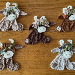 May include: Five crocheted cow ornaments with floral headbands. The cows are brown, beige, and white with different coloured flowers.