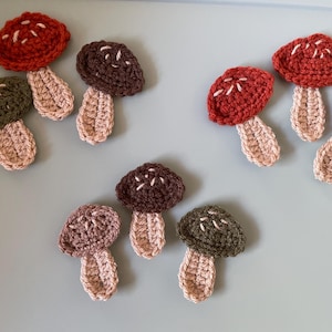 May include: A collection of crocheted mushroom ornaments in various colours, including red, brown, and green. The mushrooms have a textured, knitted appearance and are perfect for adding a whimsical touch to any space.