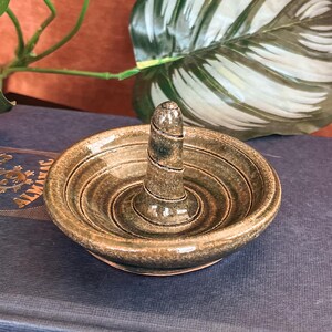 May include: A small, handcrafted ceramic ring holder in shades of green and brown. The ring holder has a central post and a shallow dish with concentric circles. It sits atop a stack of books, with a leafy plant in the background.