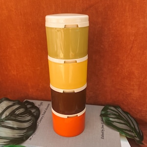 May include: A cylindrical stackable container set featuring four individual compartments in olive green, yellow, dark brown, and orange, topped with a white lid. The containers are smooth and glossy, stacked vertically to form a single tower.