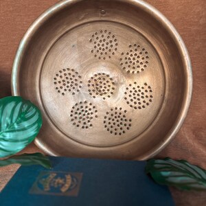 May include: A vintage copper colander with a circular design and numerous small holes for draining. The colander has a rounded rim and a small handle. The colander is a warm copper color.