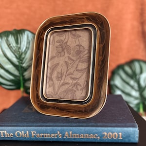 May include: A rectangular, gold-toned picture frame with a decorative border. The frame holds a floral patterned print. The frame is resting on a blue book with the title "The Old Farmer's Almanac, 2001".