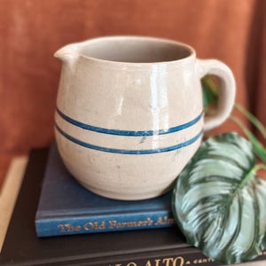 May include: A cream-colored ceramic pitcher with two horizontal blue stripes. The pitcher has a handle and a spout. It is sitting on top of a stack of books, including one titled "The Old Farmer's Almanac."