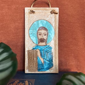 May include: A rectangular ceramic wall plaque featuring a hand-painted figure with a brown beard and hair, wearing a blue tunic against a turquoise circular halo. The piece has a light beige speckled glaze and a brown leather cord for hanging.