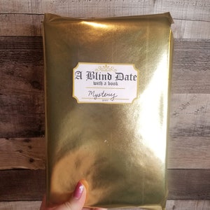 May include: A gold-wrapped book with a white label that reads "A Blind Date with a book" and "Mystery" genre.
