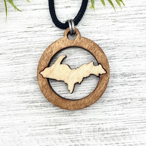 May include: A wooden necklace with a circular pendant featuring a cutout of the Upper Peninsula of Michigan. The pendant is made of light brown wood with a darker brown border. The necklace is on a black cord.