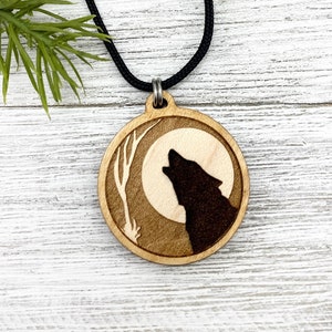 Può includere: Una collana con ciondolo in legno e un cordoncino nero. Il ciondolo è rotondo e presenta una silhouette di un lupo che ulula alla luna. La luna è circondata da rami di alberi.
