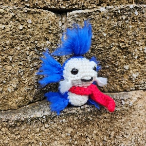 May include: A small crocheted blue and white toy with a red scarf. The toy has a fluffy blue tail and a white face with black eyes.