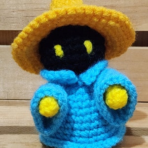 Puede incluir: Un juguete de peluche negro y azul tejido a crochet que lleva un sombrero amarillo. El juguete tiene ojos amarillos y lleva una túnica azul.