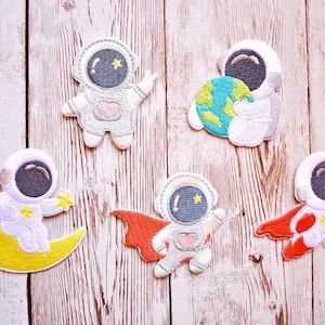 May include: Five embroidered iron-on patches featuring astronauts in various poses. The patches are white with black and grey details. One astronaut is holding a planet, one is flying with a red cape, one is holding a star, and two are on a crescent moon.