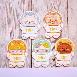 May include: Five embroidered patches featuring different animals dressed as astronauts. The animals are a white bear, a brown pig, a brown dog, a calico cat, and a brown bear. Each patch has a yellow background and a white outline.