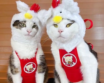 May include: Two white and tabby cats wearing red chicken costumes with a white fluffy chicken head and a red bib with a cartoon chicken eating a drumstick. The cats are sitting on a brown surface.