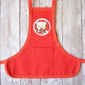 May include: Red cotton apron with a white pocket and a cartoon cat eating a chicken leg in a white circle on the front.