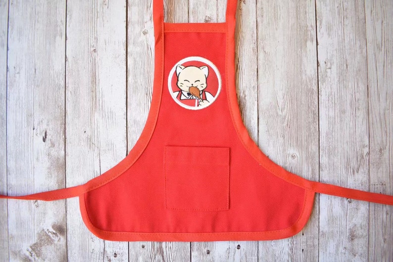 May include: Red cotton apron with a white pocket and a cartoon cat eating a chicken leg in a white circle on the front.
