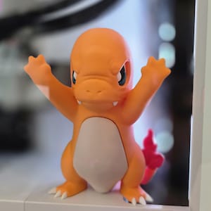 May include: An orange Charmander figurine with its arms raised. The figurine has a white belly, blue eyes, and a red flame on its tail. The figure is standing on a white surface.