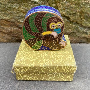 May include: A decorative trinket box featuring a colorful monkey design. The monkey is depicted in brown, green, blue, and yellow enamel, sitting atop a gold-colored box with a floral pattern. The box is likely made of metal and is a unique decorative item.