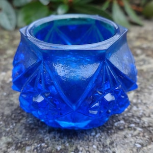 Puede incluir: Un pequeño tarro de vidrio azul cobalto con un diseño geométrico. El tarro tiene una forma hexagonal y una superficie lisa y pulida.