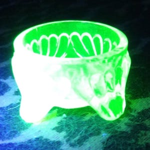 Puede incluir: Un cuenco de vidrio verde brillante, sostenido por tres pequeñas patas. El borde del cuenco presenta un diseño festoneado, y el exterior tiene un patrón texturizado. El vidrio es translúcido, mostrando un tono verde vibrante, casi neón.