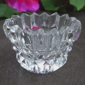 May include: A clear, faceted glass bowl with a decorative, geometric design. The bowl has a wide, open top and a multi-layered, diamond-cut pattern. The bowl is likely used for decorative purposes or as a small dish. The bowl is sitting on a dark surface.