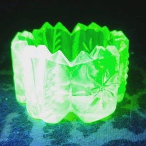 May include: A glowing green glass votive candle holder with a geometric design. The clear glass is etched with floral and leaf patterns. The candle holder has a scalloped top edge and a square base.