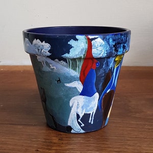 May include: A hand-painted terracotta flower pot featuring a blue, white and red landscape scene. The scene depicts a white horse, a figure in a red robe and a figure in a blue robe.