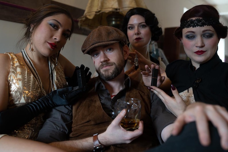 Puede incluir: Un grupo de personas vestidas con atuendos de la d&eacute;cada de 1920. Un hombre con un fedora marr&oacute;n y un chaleco sostiene un vaso de whisky. Una mujer con un vestido negro sostiene un l&aacute;piz labial. Otra mujer con un vestido de lentejuelas doradas lleva guantes negros.