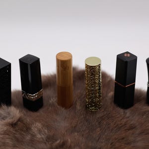 Puede incluir: Seis tubos de l&aacute;piz labial diferentes con varios dise&ntilde;os y colores. Los tubos est&aacute;n dispuestos en una fila sobre una superficie de piel marr&oacute;n. Los tubos est&aacute;n hechos de negro, oro y madera.