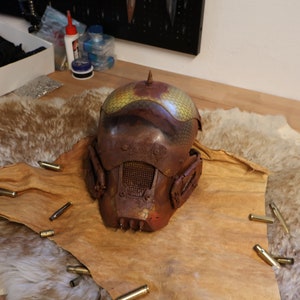Könnte beinhalten: Ein rostiger, brauner Helm mit einem gelben und grünen Muster auf der Oberseite. Der Helm hat eine Gesichtsmaske aus Metallgitter und einen Dorn oben. Der Helm liegt auf einem braunen Fellteppich mit mehreren Patronenhülsen, die um ihn herum verstreut sind.