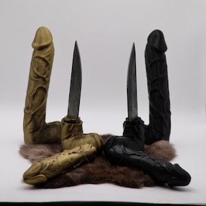 Könnte beinhalten: Vier dekorative Skulpturen, zwei goldfarbene und zwei schwarze, jeweils mit einer phallischen Form mit eingesetzter Messerklinge. Die Skulpturen ruhen auf einem Bett aus braunem Fell vor weißem Hintergrund.