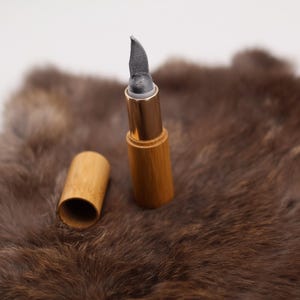 Puede incluir: Un tubo de madera con una punta gris puntiaguda y una banda de metal dorado. El tubo est&aacute; sobre una superficie de piel marr&oacute;n.