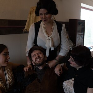 Puede incluir: Un grupo de cuatro personas en una habitaci&oacute;n con poca luz. Una mujer con una blusa blanca y un chaleco negro est&aacute; de pie sobre un hombre sentado en un sof&aacute;. El hombre est&aacute; mirando hacia arriba con una expresi&oacute;n de sorpresa. Otras dos mujeres est&aacute;n sentadas en el sof&aacute;, una a cada lado del hombre. La mujer de la izquierda lleva un vestido de lentejuelas doradas y la mujer de la derecha lleva un vestido negro.