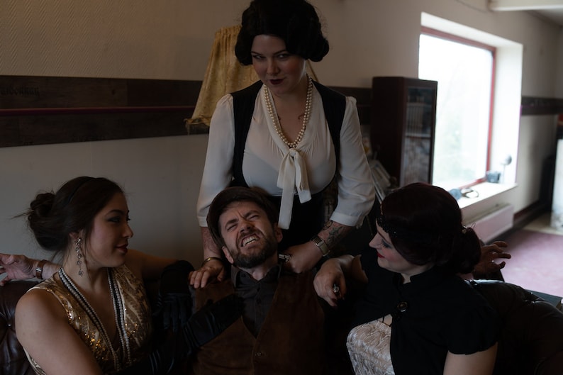 Puede incluir: Un grupo de cuatro personas en una habitaci&oacute;n con poca luz. Una mujer con una blusa blanca y un chaleco negro est&aacute; de pie sobre un hombre sentado en un sof&aacute;. El hombre est&aacute; mirando hacia arriba con una expresi&oacute;n de sorpresa. Otras dos mujeres est&aacute;n sentadas en el sof&aacute;, una a cada lado del hombre. La mujer de la izquierda lleva un vestido de lentejuelas doradas y la mujer de la derecha lleva un vestido negro.