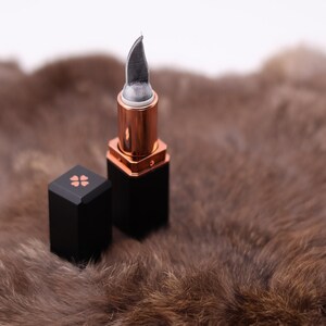 Puede incluir: Un tubo de l&aacute;piz labial negro con una cuchilla plateada que se extiende desde la parte superior. El l&aacute;piz labial est&aacute; sobre una superficie de piel marr&oacute;n.