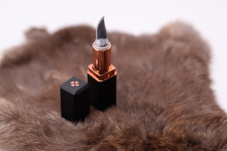 Puede incluir: Un tubo de l&aacute;piz labial negro con una cuchilla plateada que se extiende desde la parte superior. El l&aacute;piz labial est&aacute; sobre una superficie de piel marr&oacute;n.