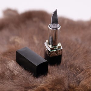 Puede incluir: Un pintalabios plateado con una punta afilada, sobre una superficie de piel marr&oacute;n. El pintalabios est&aacute; cerrado y tiene una tapa negra.